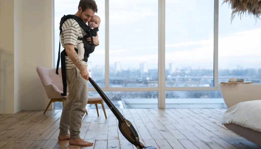 Homem usando aspirador portátil Electrolux vertical enquanto carrega bebê - praticidade na limpeza doméstica