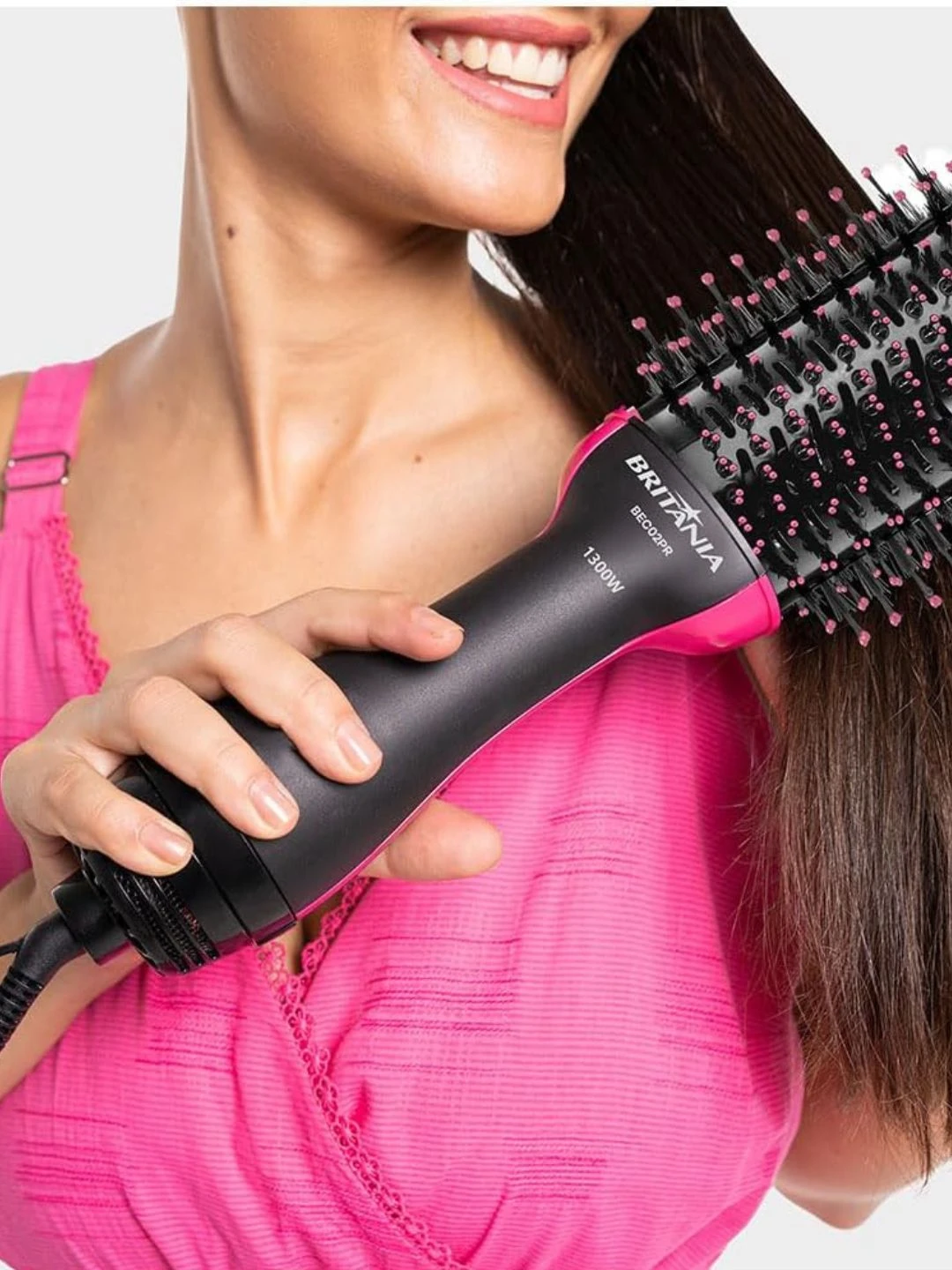 Mulher jovem sorrindo enquanto alisa o cabelo com a escova secadora Britânia Soft 4 em 1, em fundo claro.